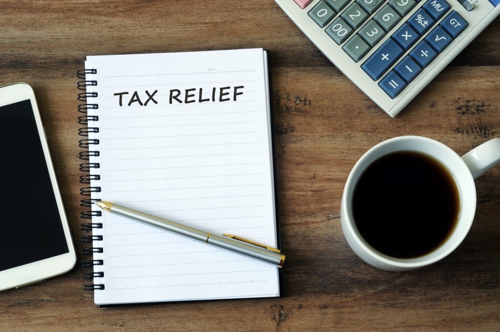 Financial concept - Text Tax relief on a notepad with smart phone, pen, coffee and calculator on a wooden table. Financial concept - Text Tax relief on a notepad with smart phone, pen, coffee and calculator on a wooden table.